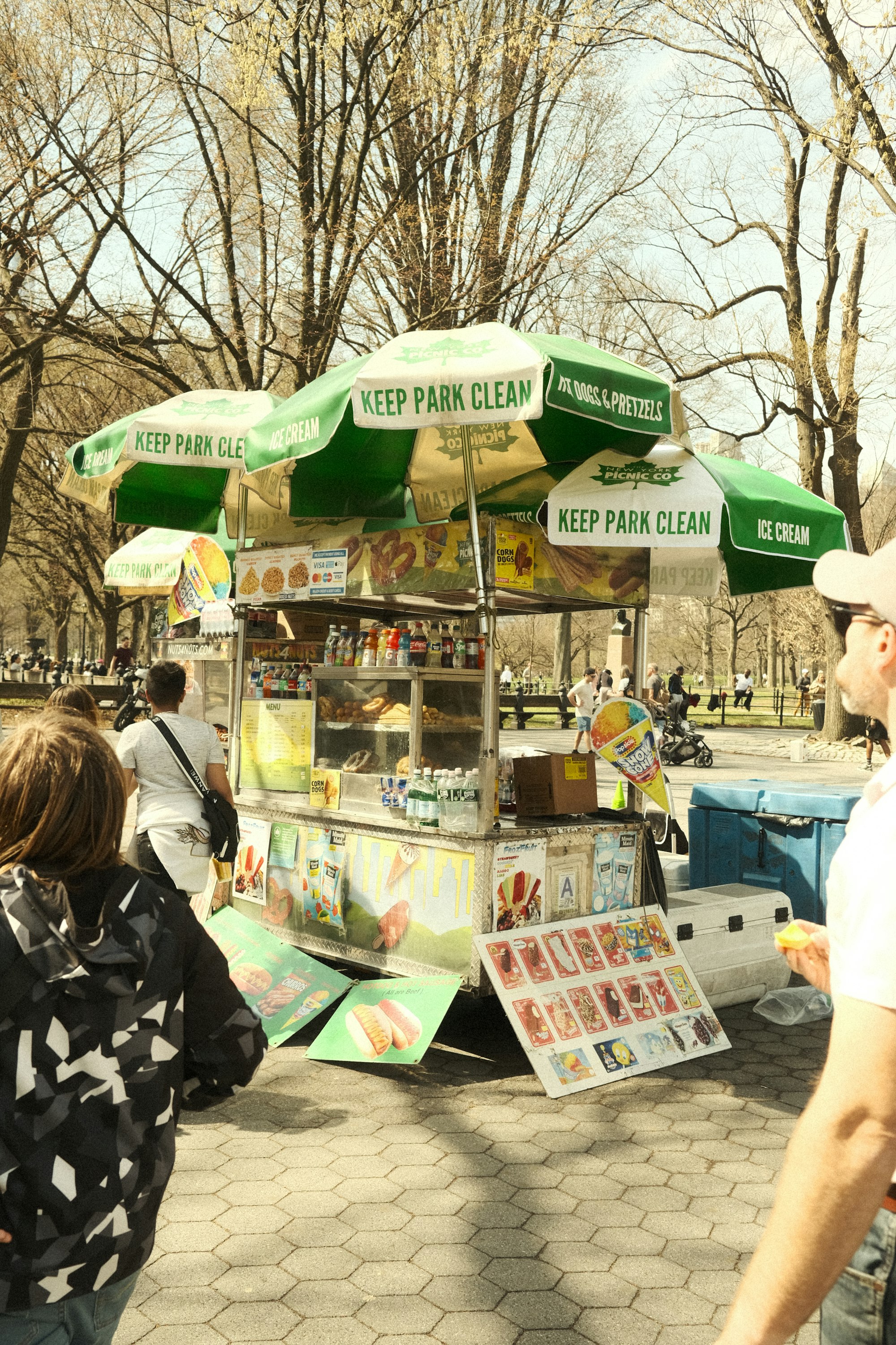 A red hot dog cart with a striped umbrella set up on a city sidewalk